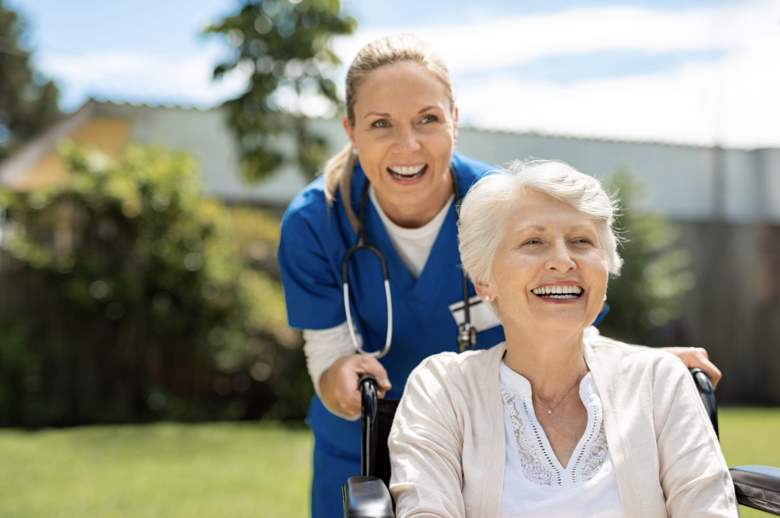 Happy nurse and elderly woman sitting in wheelchair enjoying outdoor treatment session. Beautiful nurse with laughing senior woman in wheelchair at outdoor park. Smiling disabled old lady in wheelchair at park.